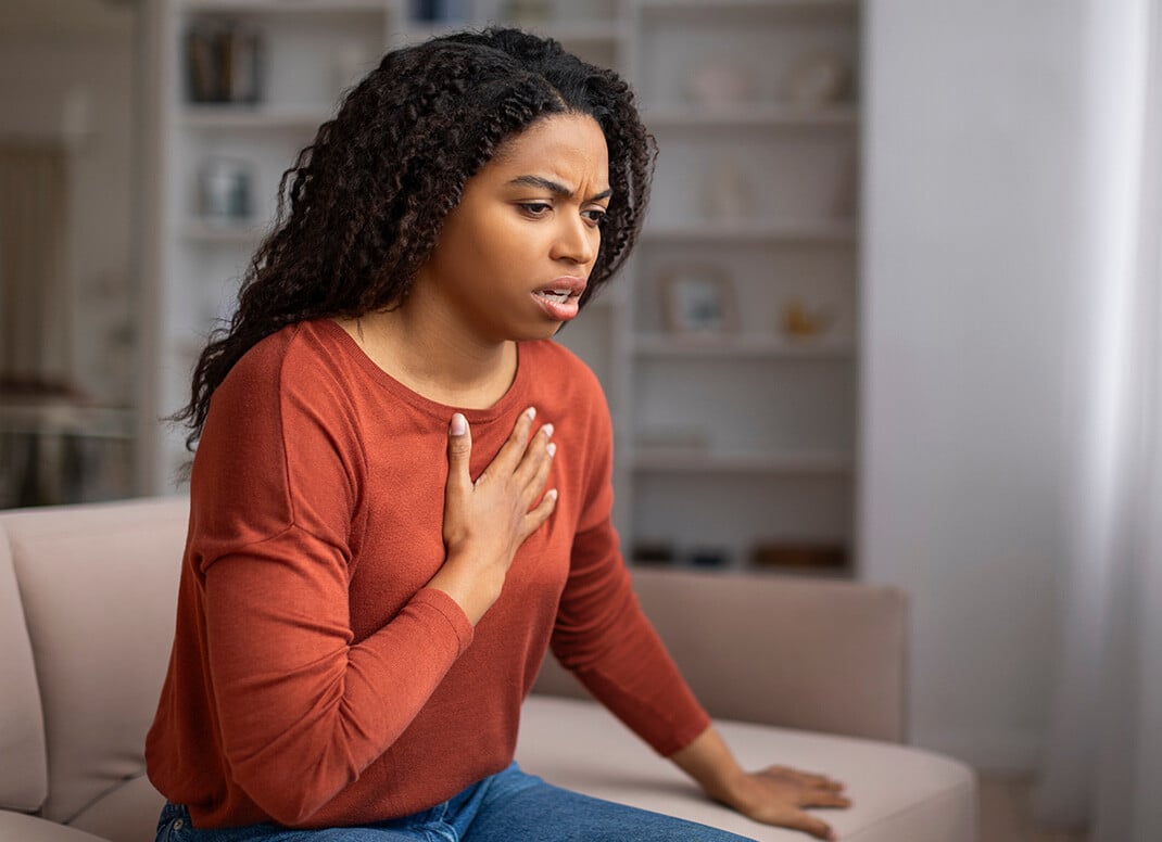 A concerned woman sitting on a couch holds her chest with one hand
